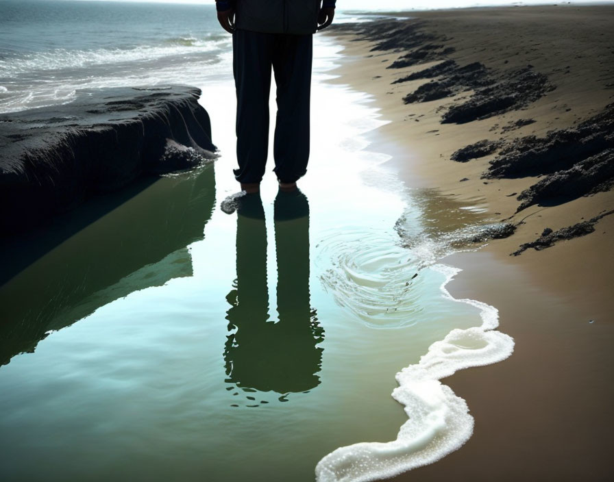 Person standing at water's edge on sandy beach with reflection in calm water.