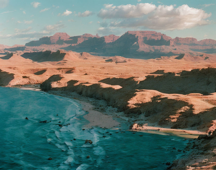 Tranquil beach meets rugged desert landscape with mesas under soft blue sky