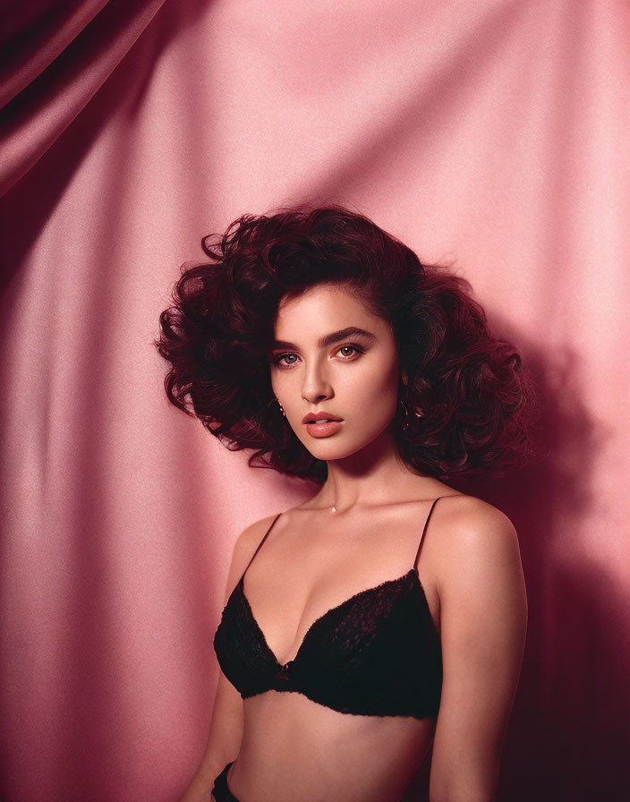 Portrait of woman with curly hair, piercing eyes, dark lipstick, posing in black lace against pink backdrop