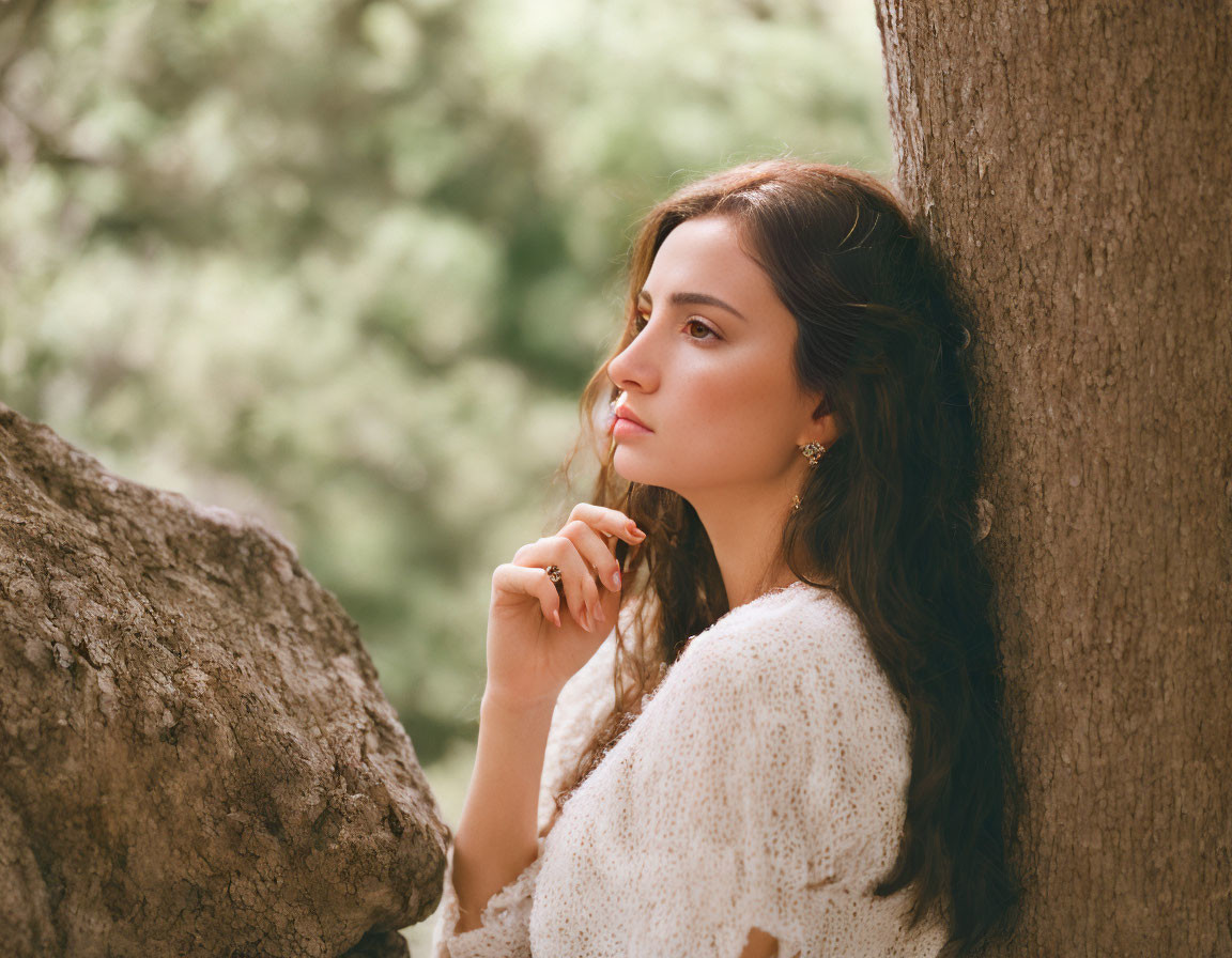 Dark-haired woman in white outfit leaning against tree in serene nature scene