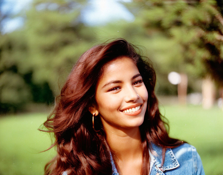 Smiling woman with long hair in denim jacket outdoors among green trees.