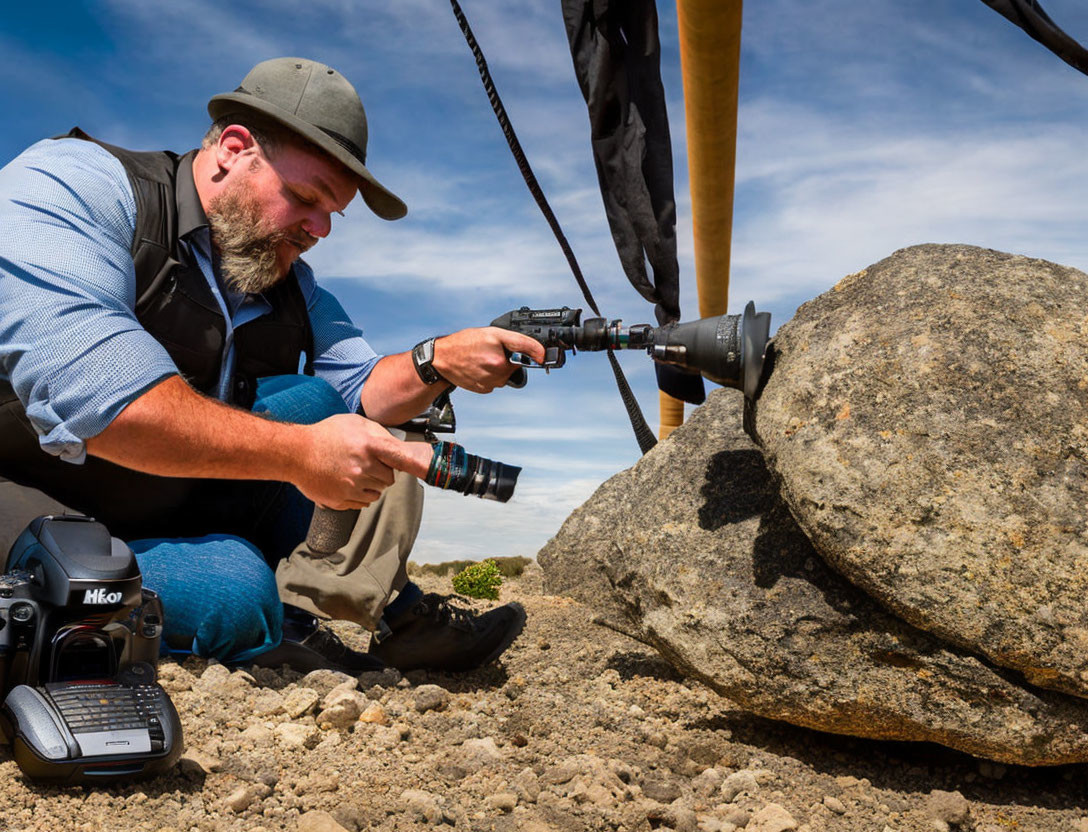 Photographer in hat and vest capturing subject behind rock with camera gear.