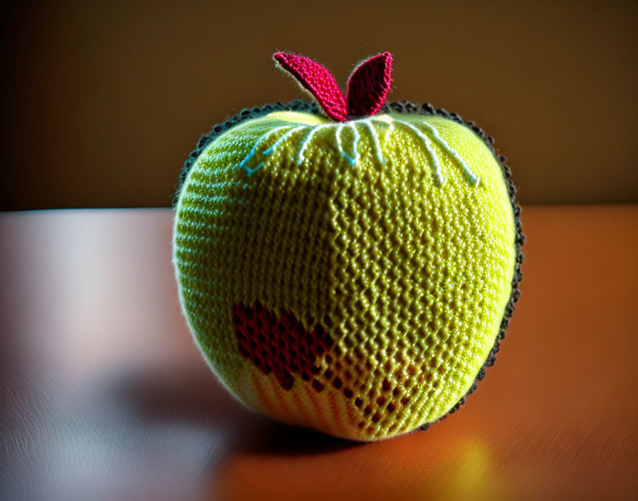 Green apple crocheted item with red leaf on wooden surface under soft lighting