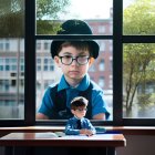 Contemplative boy in blue suit and hat seen through window with miniature version on desk