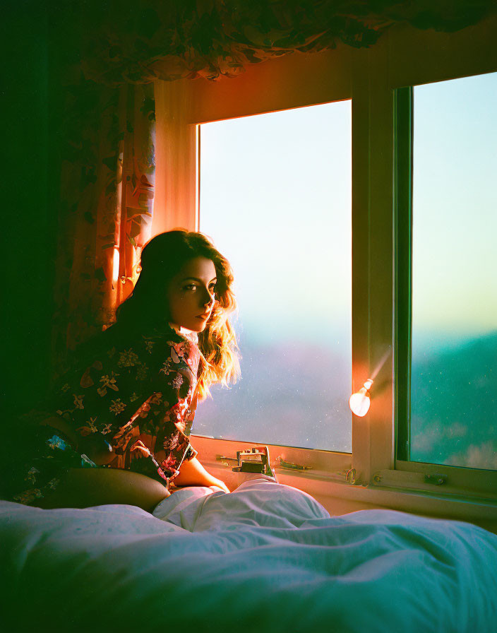 Person in floral dress by window at sunset with camera in warm, well-lit room