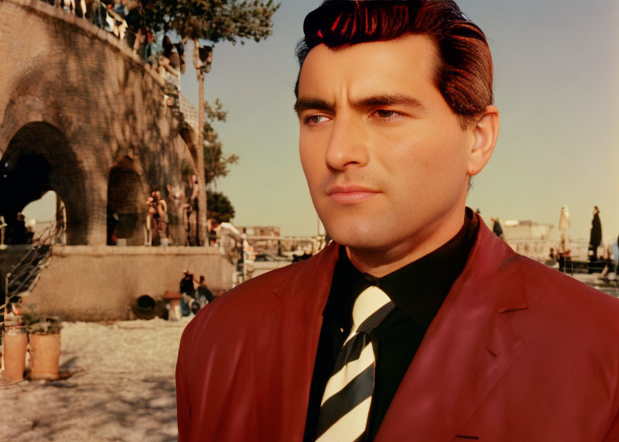 Dark-haired man in red jacket gazes pensively by bridge in vintage setting