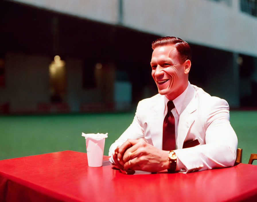 Smiling man in white suit at red table with takeout cup on green background