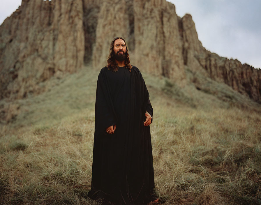 Bearded Figure in Black Robe on Grass Field with Cliff