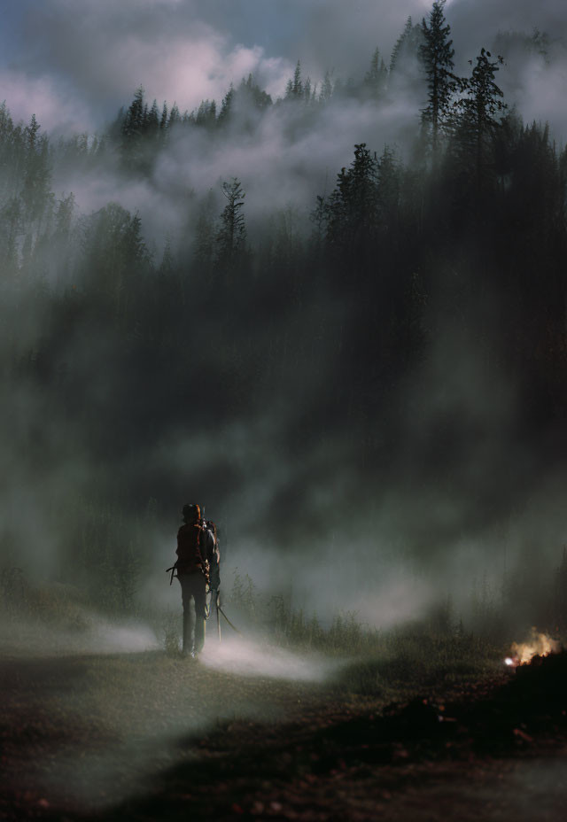 Hiker walking on misty forest path at dusk with campfire