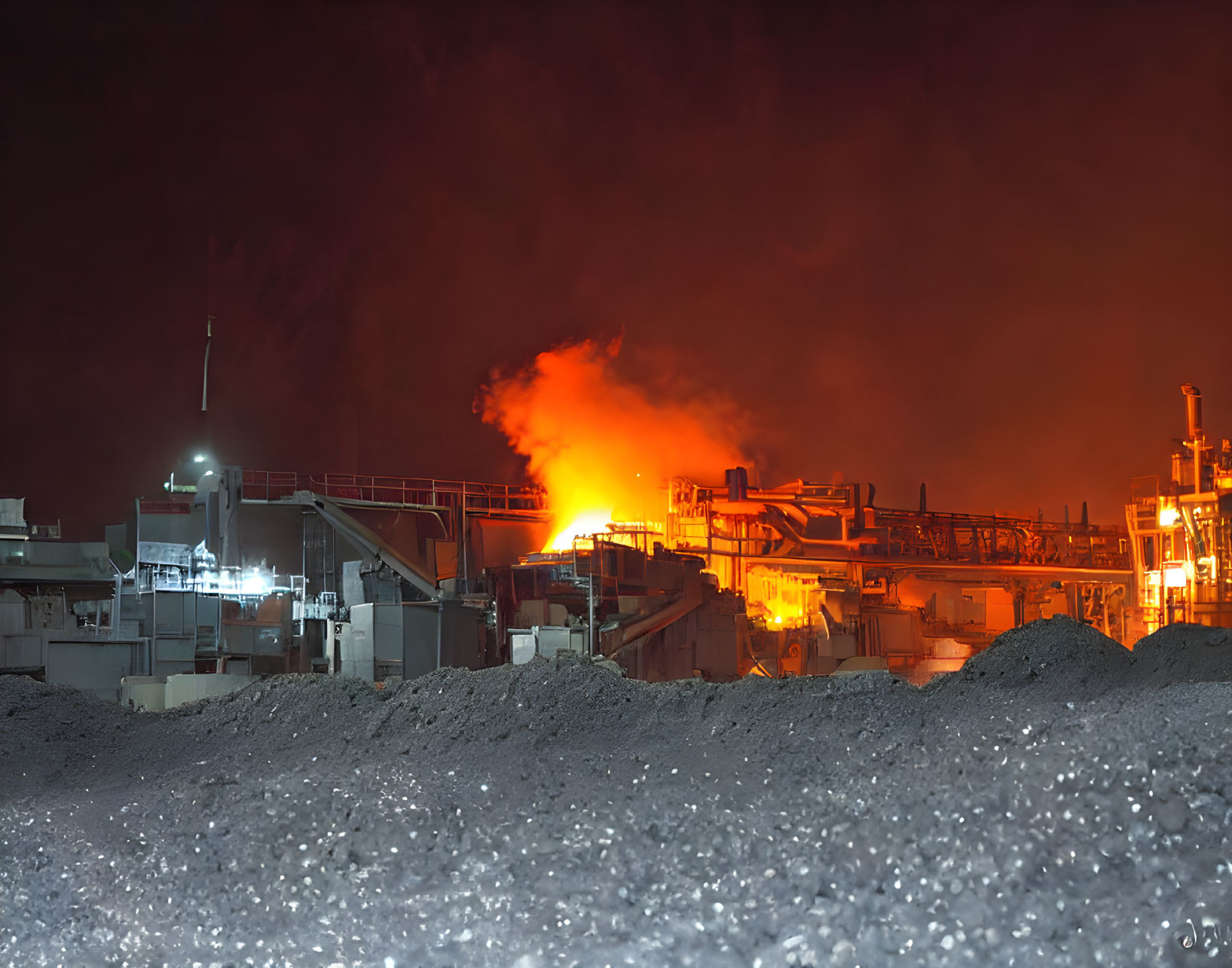 Industrial facility at night with fiery glow against dark sky & raw material heaps