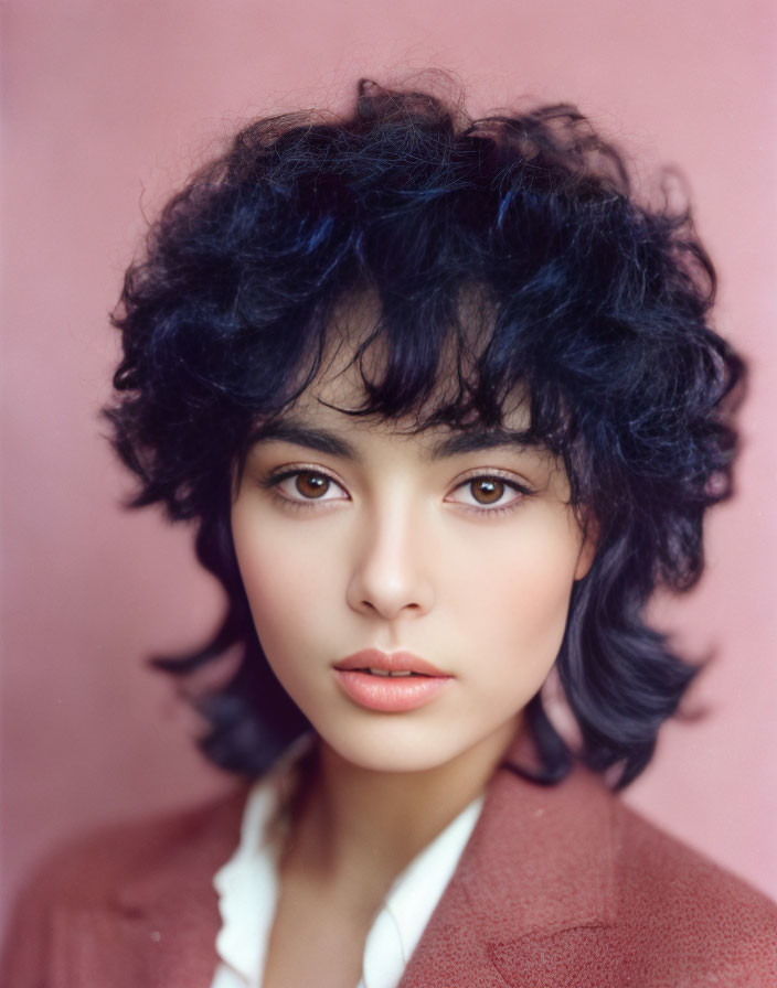 Portrait of person with voluminous curly black hair, fair skin, white shirt, and red blazer