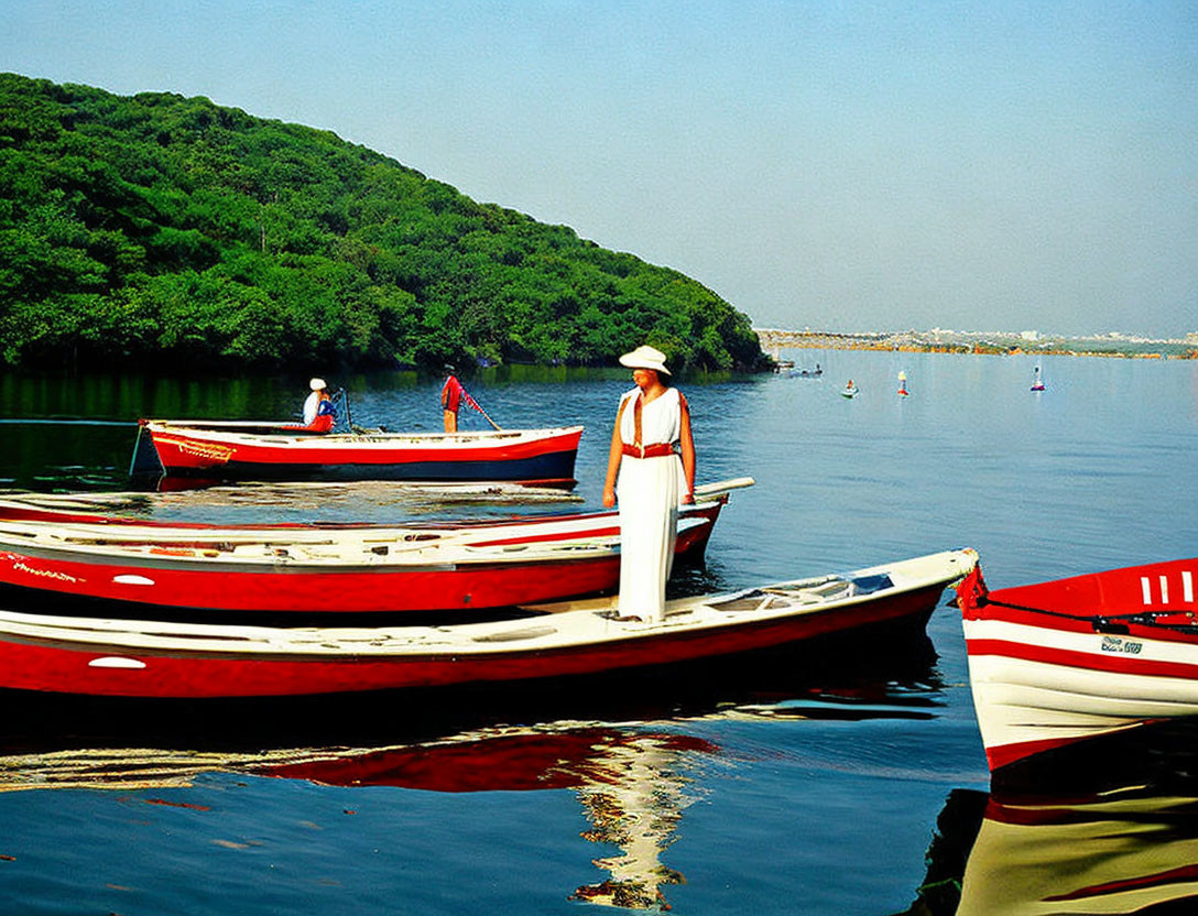 Person in White Dress and Hat by Red Boats on Calm River