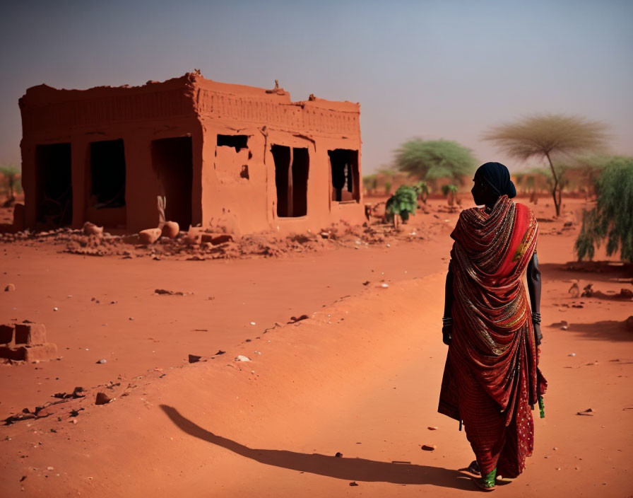 Traditional attire person in front of dilapidated red clay building in desert landscape