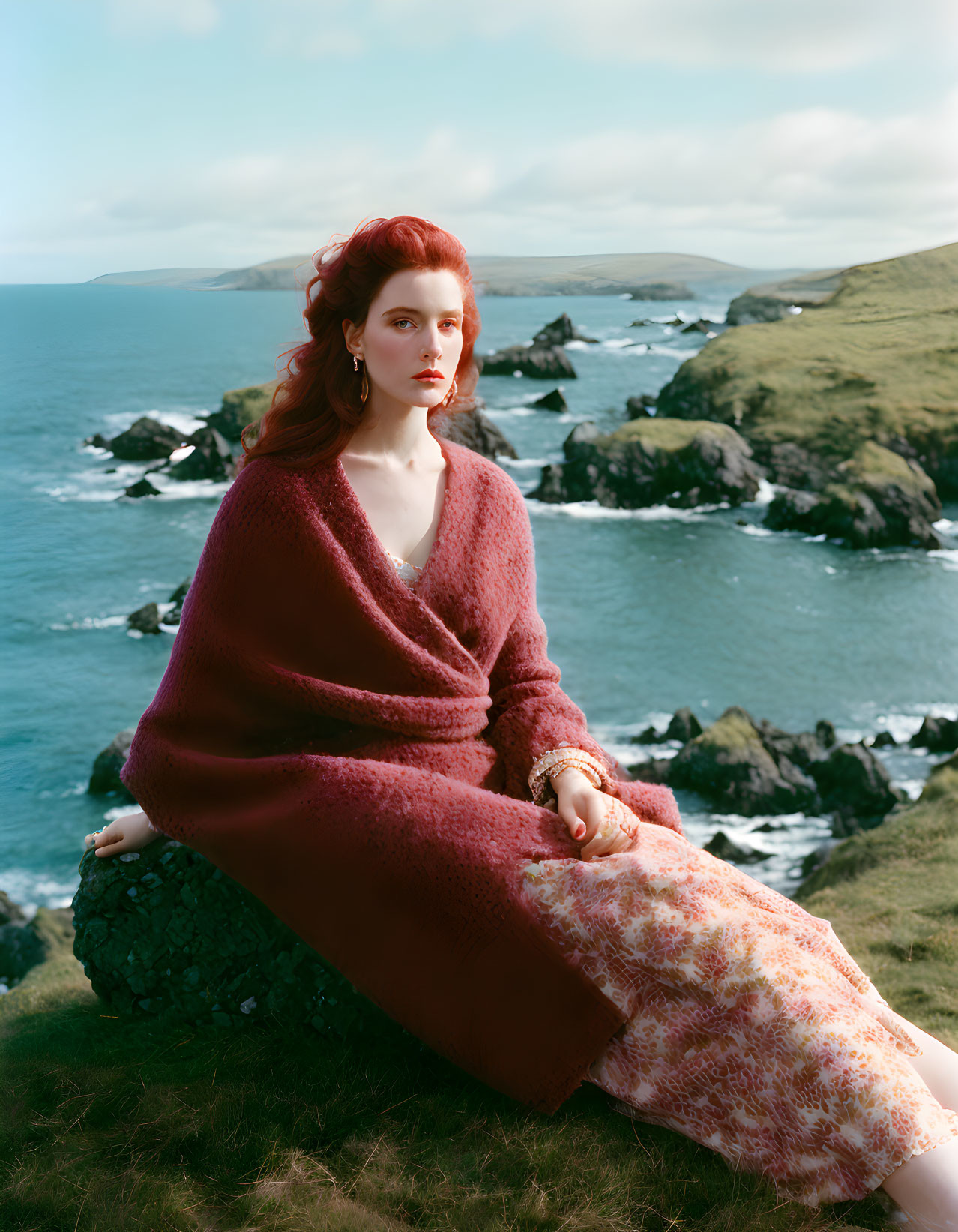 Red-haired woman in maroon wrap on grassy hill overlooking rocky coastline