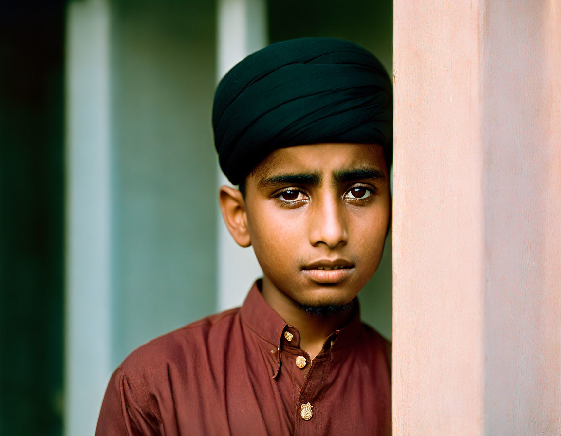 Young boy in dark turban and brown shirt near pink wall with thoughtful expression