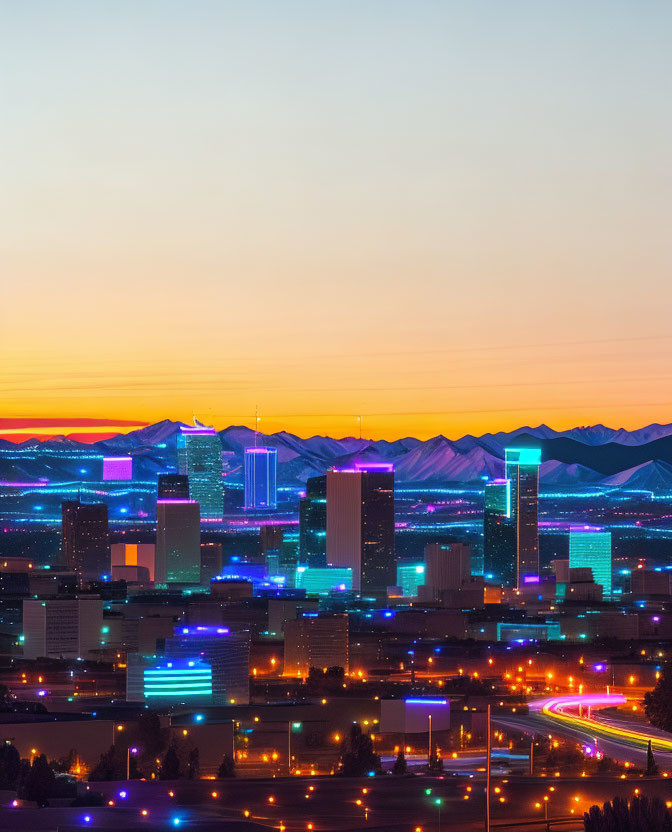 Urban skyline at twilight with glowing buildings and colorful sunset behind distant mountains.