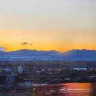 Urban skyline at twilight with glowing buildings and colorful sunset behind distant mountains.