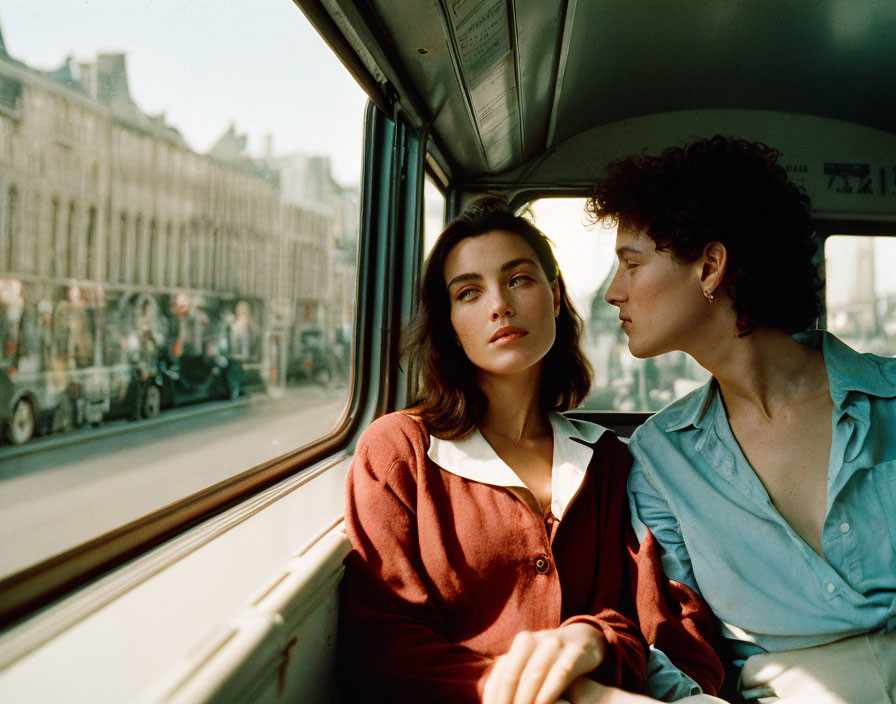 Two women on bus seat, one looking out window, city streets in soft sunlight.