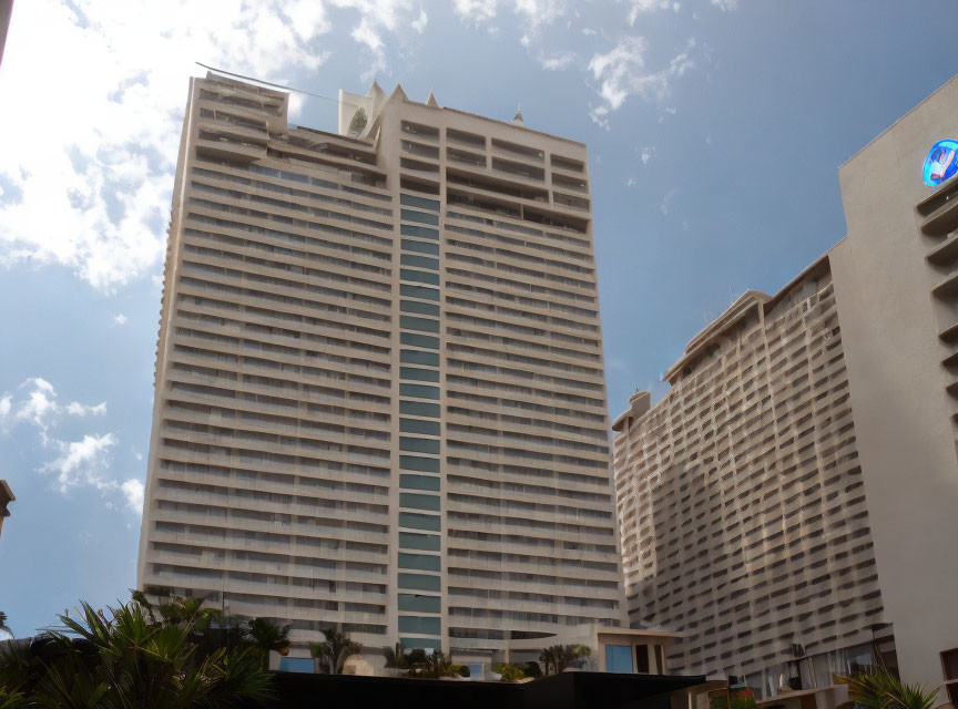 Tall hotel building with many windows under cloudy sky next to building with circular logo