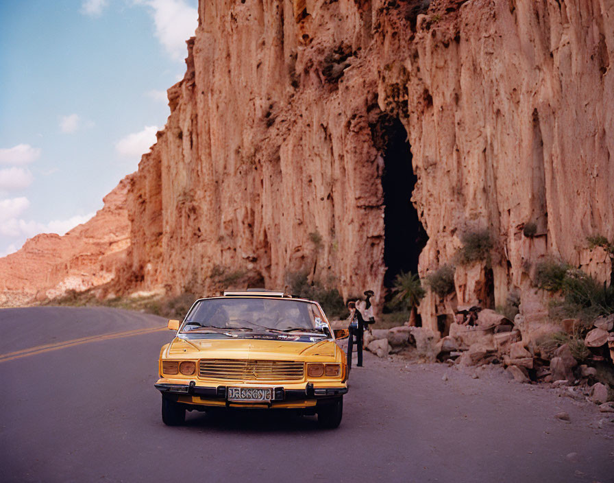 Vintage Yellow Car Parked Near Rocky Cliffs with Cave Entrance, Person and Goats Nearby