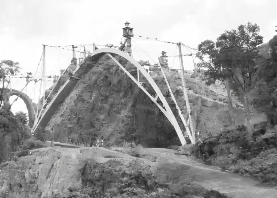 Monochrome image of arched bridge with suspension cables and lit lamps in rugged landscape.