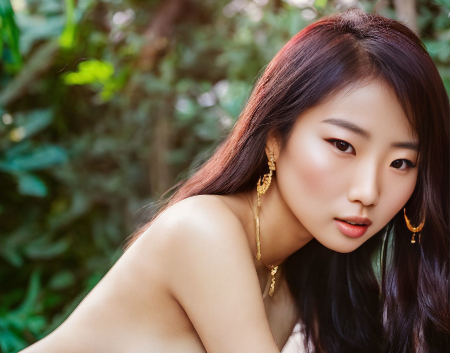 Dark-haired woman with gold earrings in front of green foliage.