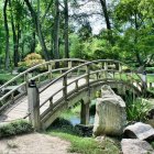 Japanese arched bridge over stream surrounded by lush greenery and pink flowering shrubs.