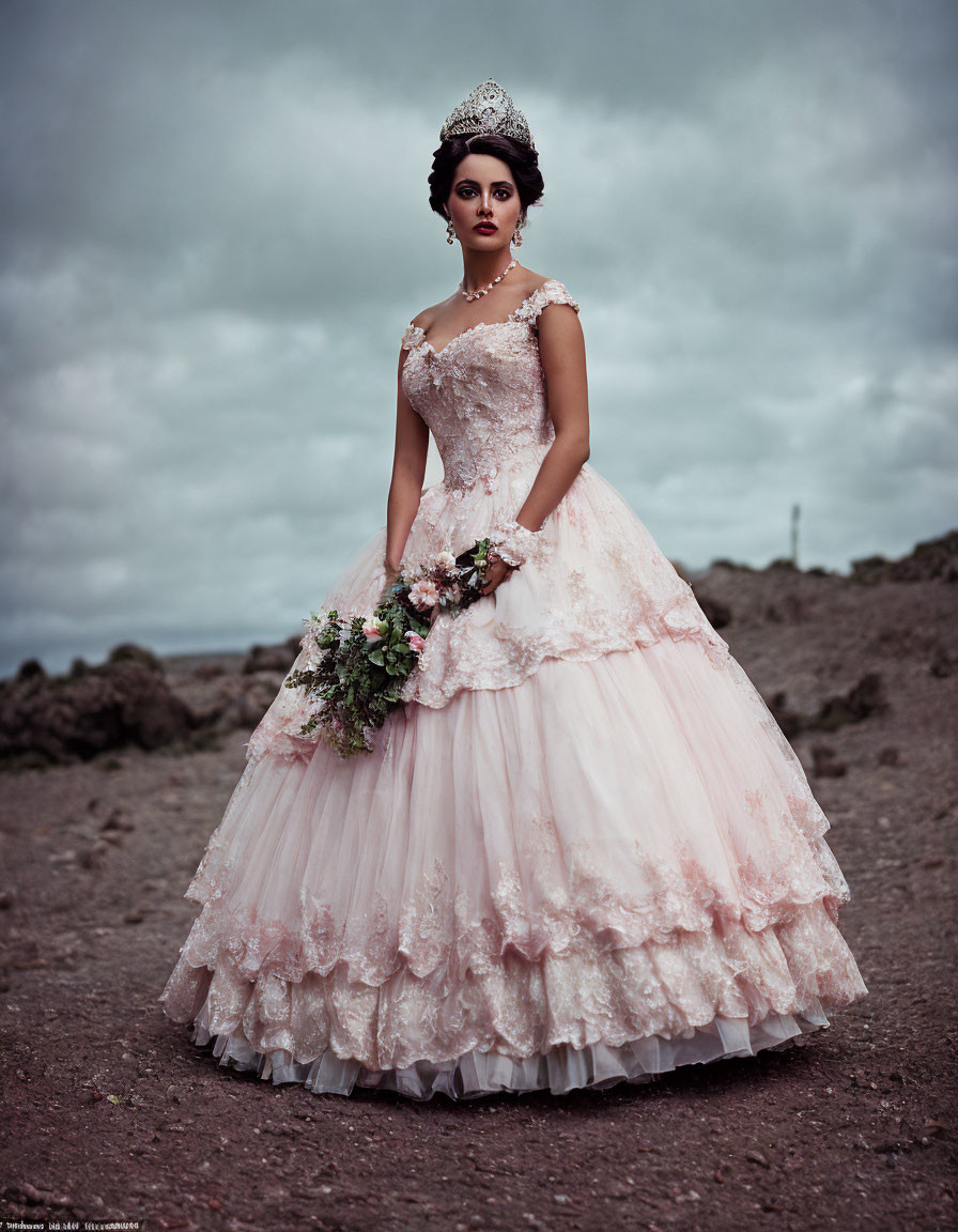 Elaborate pink gown with lace detailing and tiara against moody sky and rocky terrain