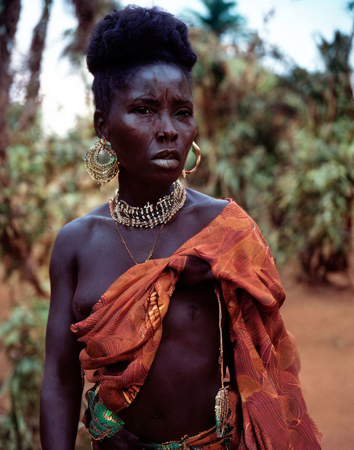 Traditional jewelry adorns woman with face markings in orange and green attire against natural backdrop