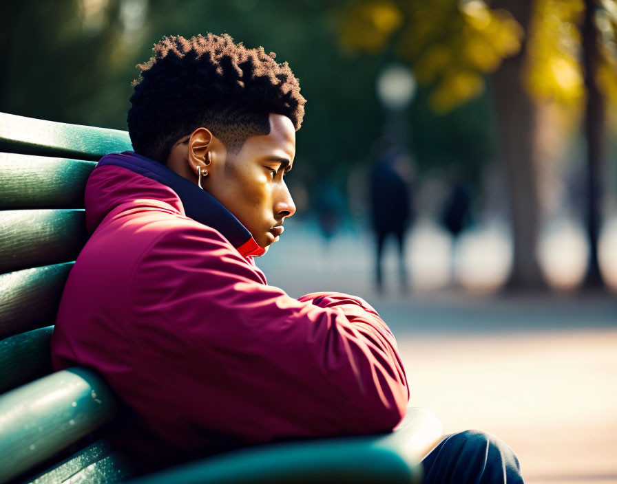 Young person with short curly hair in red jacket sitting on park bench with trees