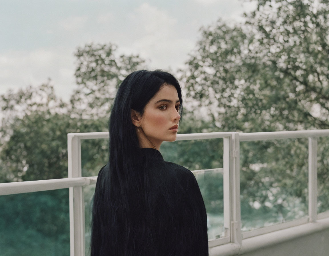 Long-haired woman gazes thoughtfully on balcony with foliage.