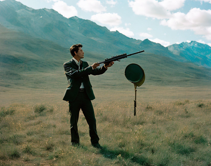 Man in suit aiming rifle outdoors with mountains and frying pan in front