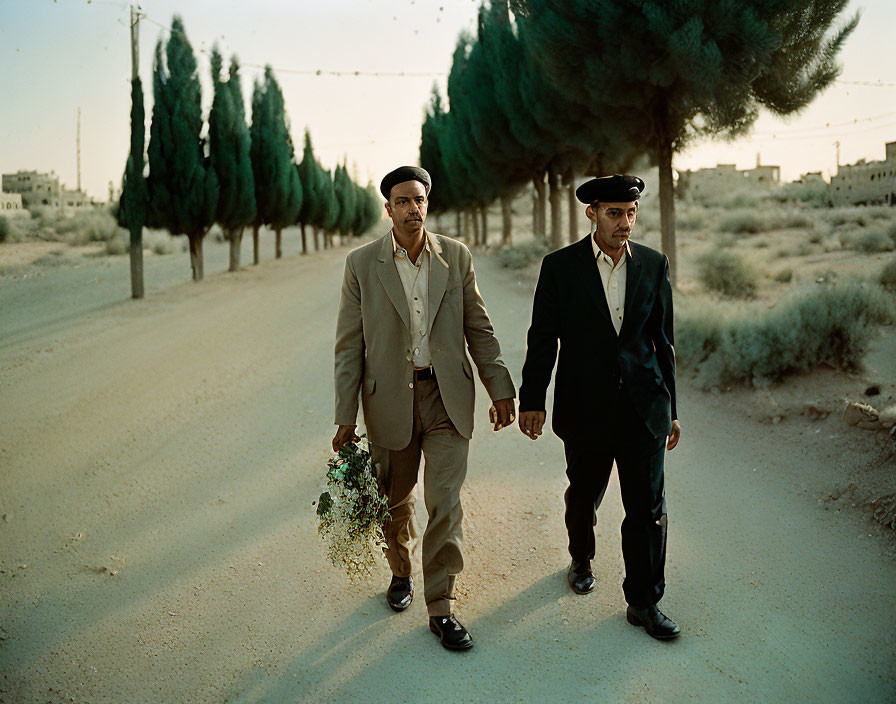 Men in suit and uniform walking on dusty road with bouquet, trees along path