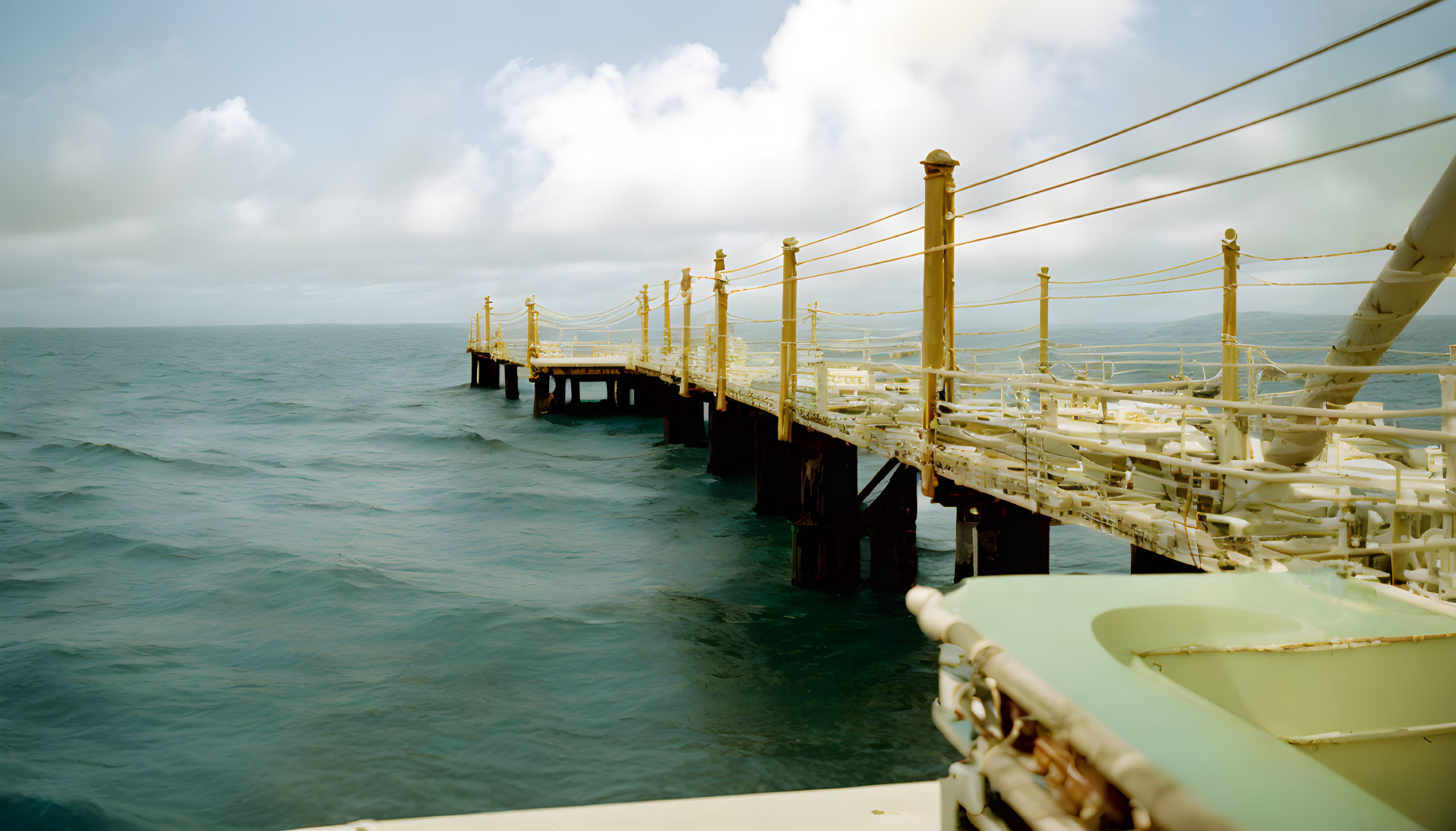 Wooden pier with rope railings extending into tranquil sea under cloudy sky.