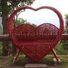 Heart-shaped bench adorned with flowers and plush pink and red cushions in a tranquil garden scene