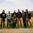 Seven men in military outfits standing in field with smoke and flying debris.