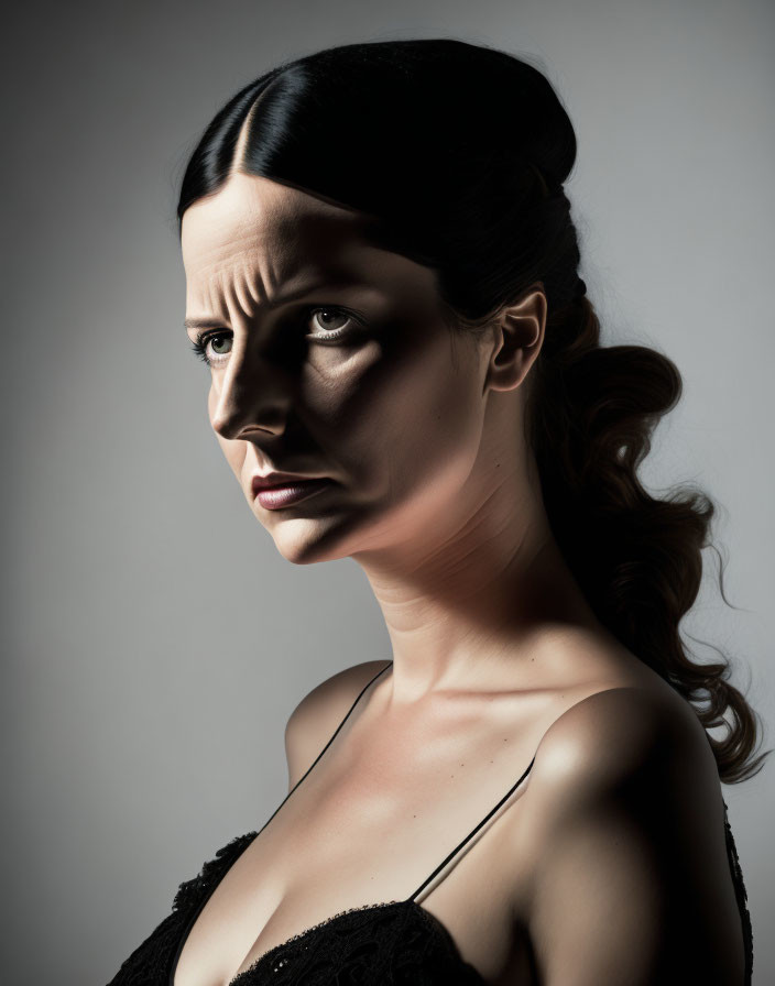 Dark-haired woman in updo and black top gazes contemplatively, half face in shadow.