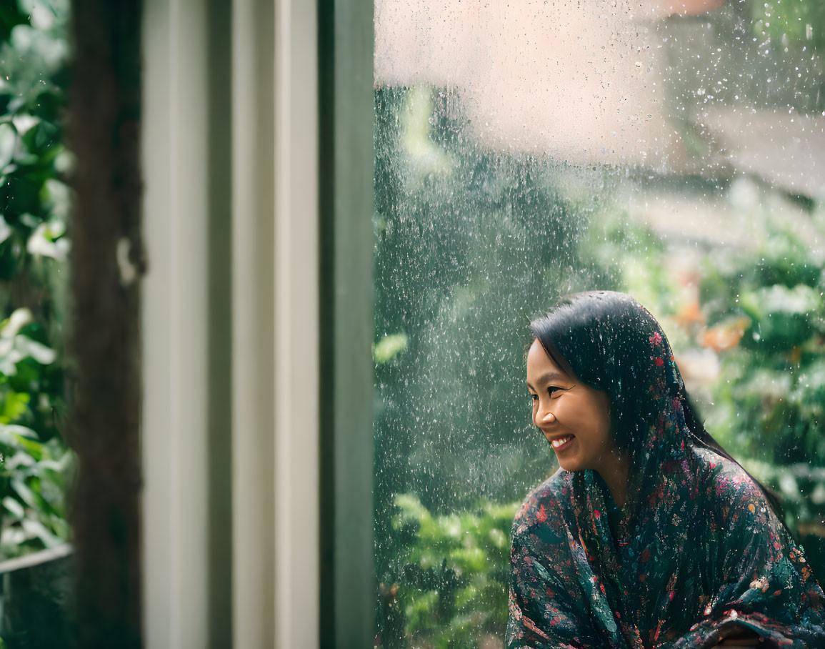 Woman in floral dress smiling by raindrop-covered window overlooking garden