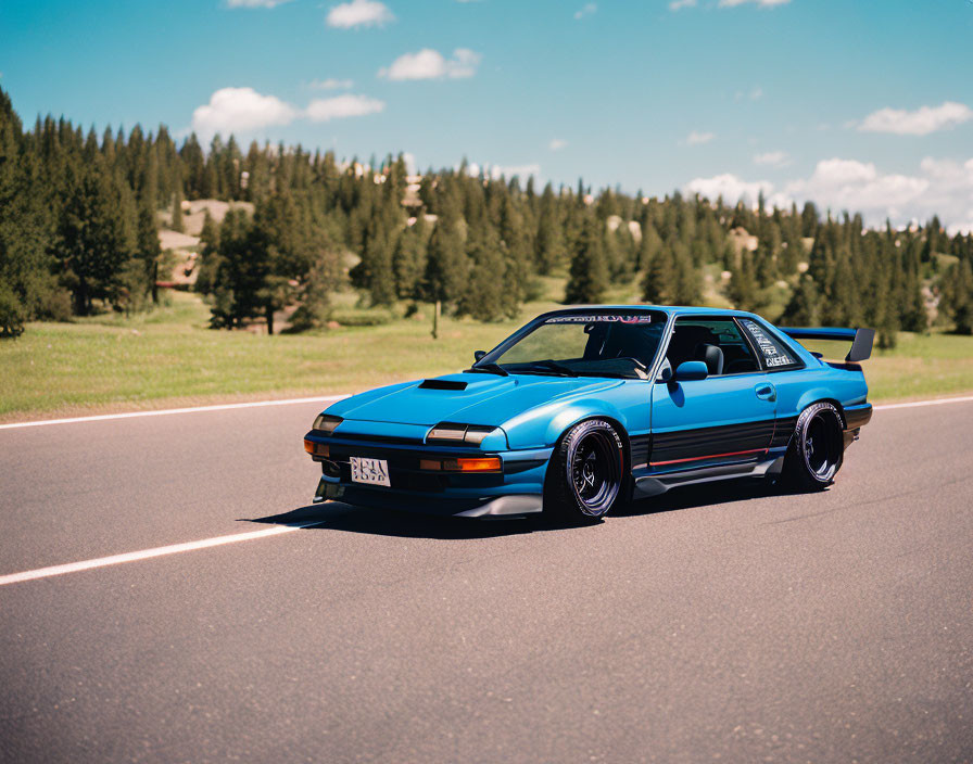 Blue Vintage Sports Car with Black Accents and Aftermarket Modifications on Asphalt Road with Green Background