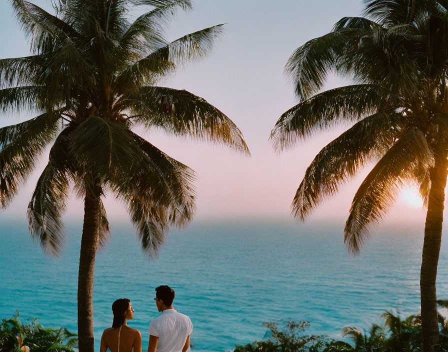 Couple standing between palm trees by ocean at sunset