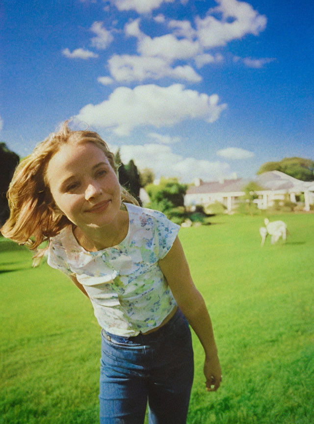 Young girl smiling in sunny meadow with house and dog.