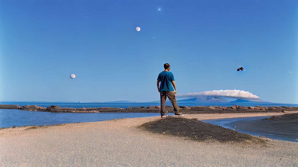 Man standing on sandy path by sea, mountains under starry sky.