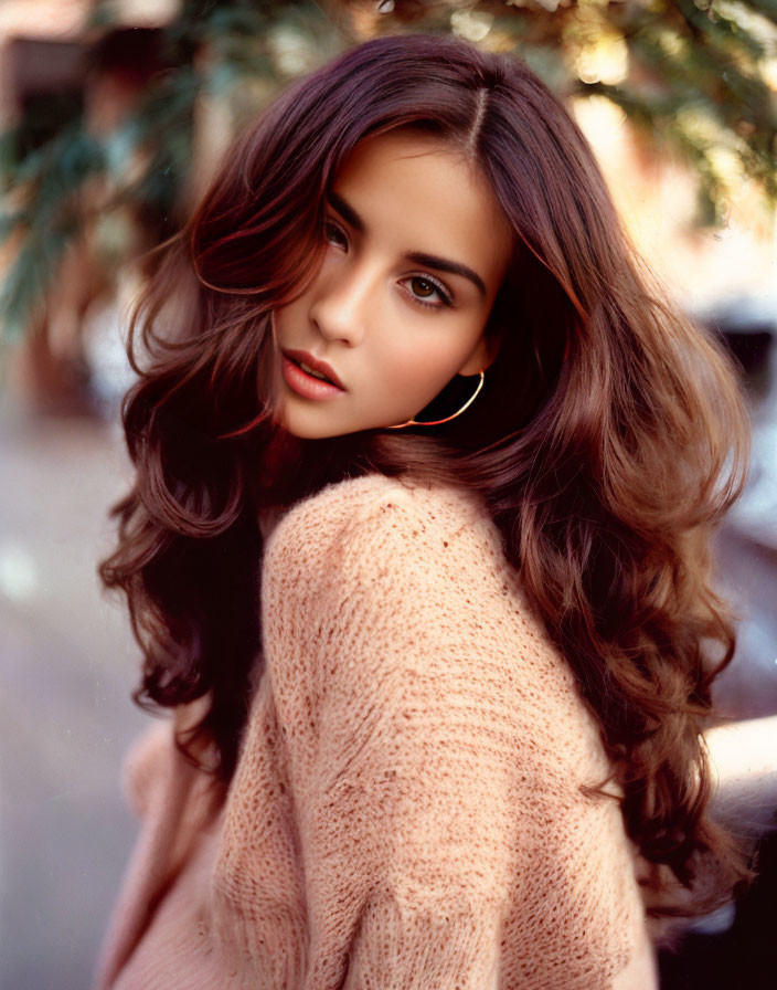 Woman with long wavy brown hair in peach sweater and hoop earrings gazes over shoulder