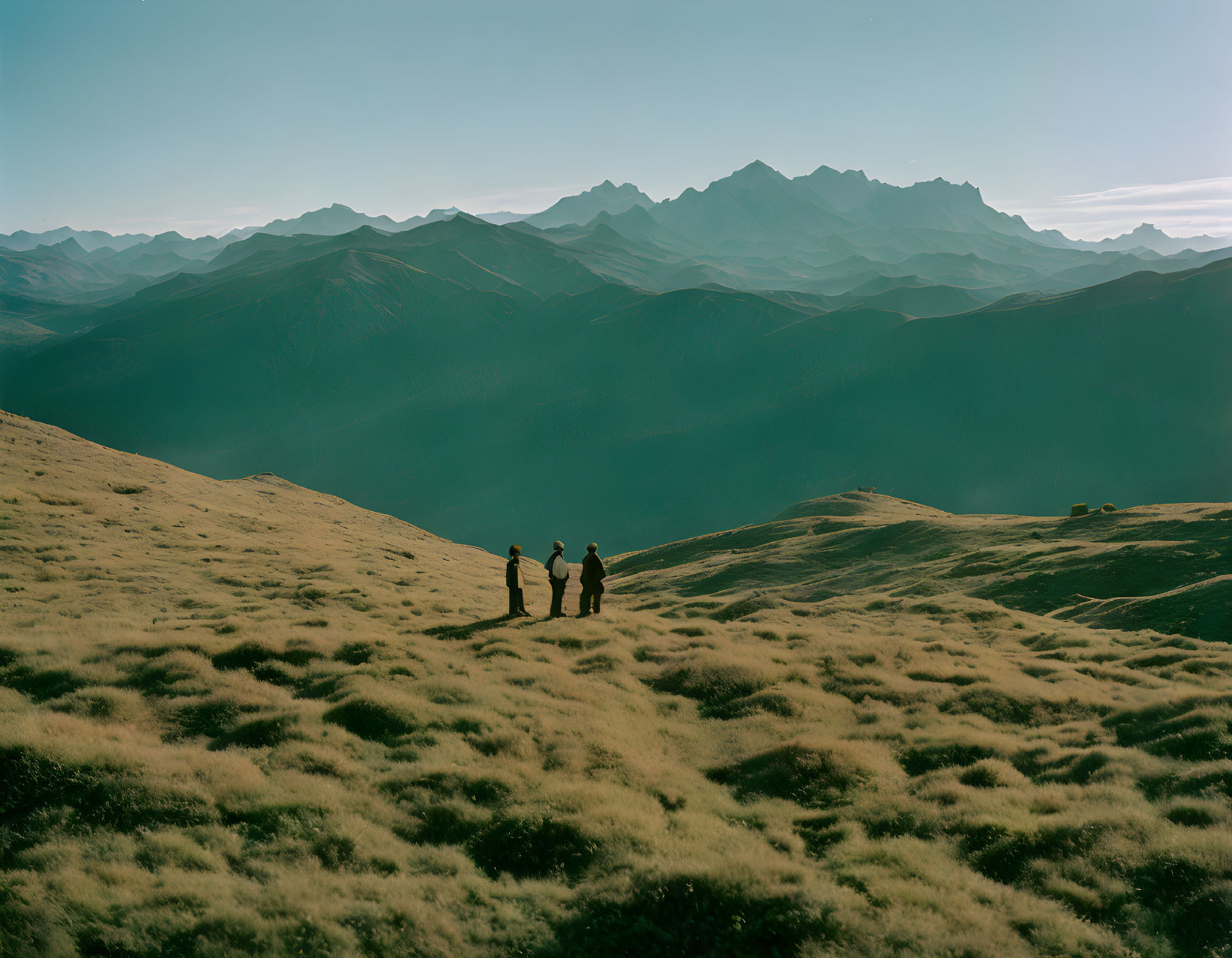 Three individuals on grassy hill with mountain backdrop under blue sky