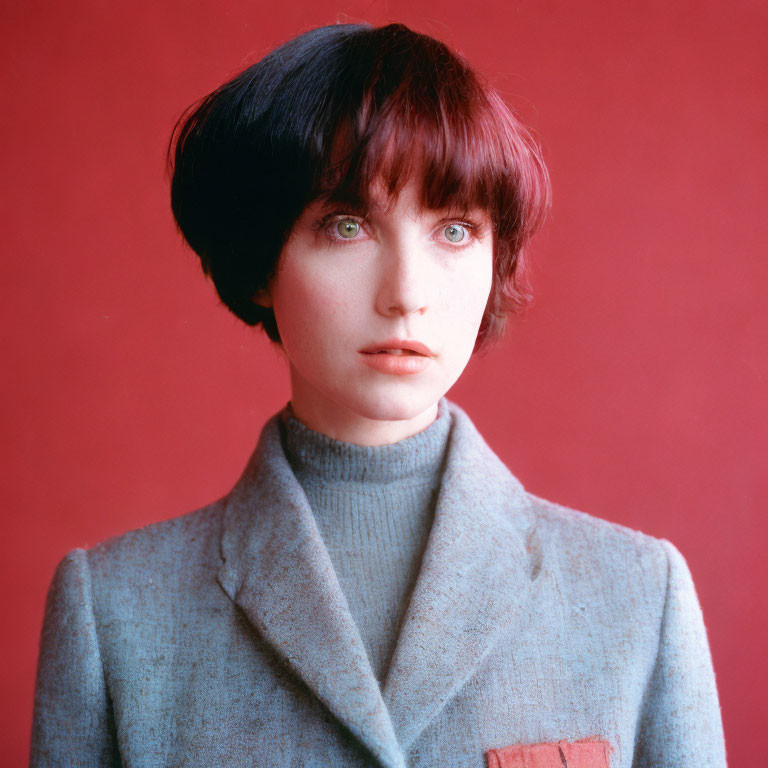 Person with Short Black Hair in Grey Turtleneck and Blazer on Red Background