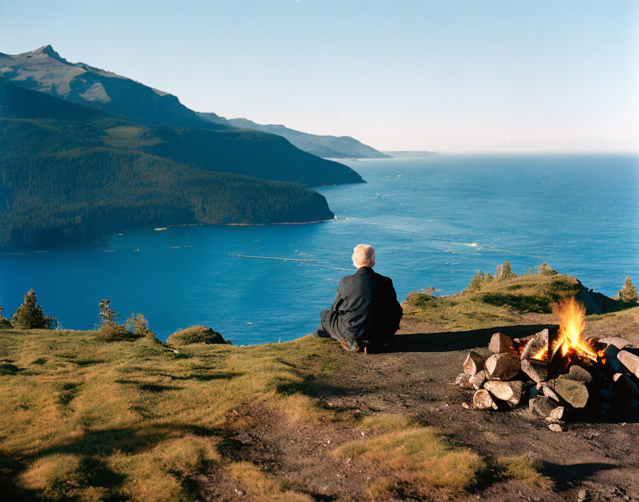 Individual enjoys campfire with coastal view and mountain backdrop.