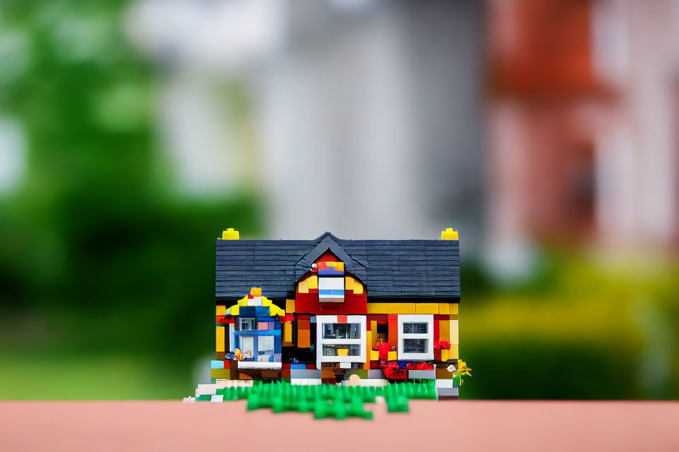 Colorful Lego House with Red Roof and Flower Details in Front of Blurred Real Houses