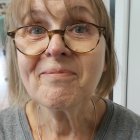 Elderly person with deep wrinkles and glasses in blue shirt by curtain