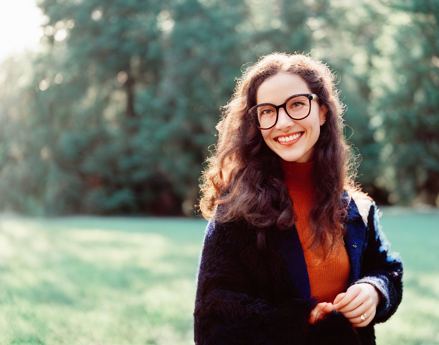 Smiling woman with glasses and curly hair in blue coat in sunlit park