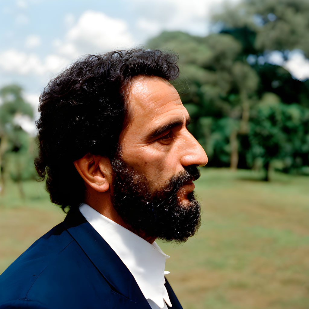 Man with Dark Hair and Full Beard in Suit Against Tree Backdrop